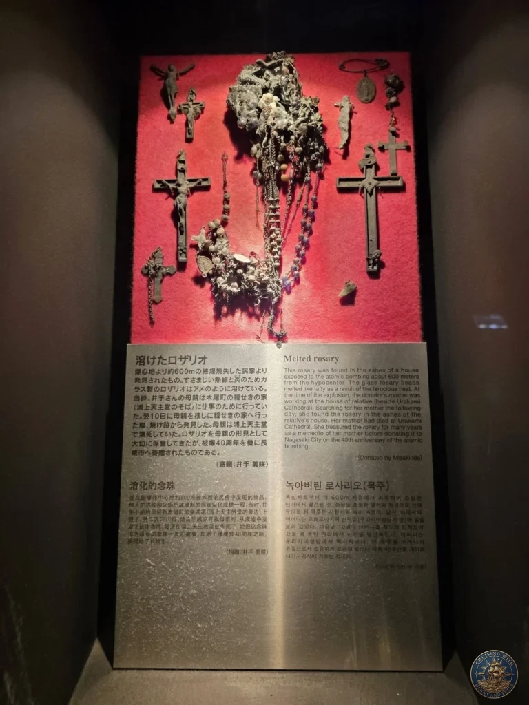 Display case at the Nagasaki Atomic Bomb Museum showing a melted rosary and several crucifixes that survived the atomic bombing. The artifacts are mounted on a red background with detailed English and Japanese explanation panels below. Featured in Nagasaki Japan cruise port guide, things to do in Nagasaki from cruise ship, and one day in Nagasaki Japan itinerary from the Westerdam. by Danny and Peter.
