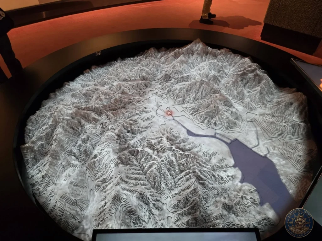 Large circular topographic relief model of Nagasaki at the Atomic Bomb Museum, showing the city's hilly terrain and harbor. A red light marks the hypocenter of the atomic bomb. Visitors can be seen observing the detailed 3D model. Featured in Nagasaki Japan cruise port guide, things to do in Nagasaki from cruise ship, and one day in Nagasaki Japan itinerary from the Westerdam. by Danny and Peter.