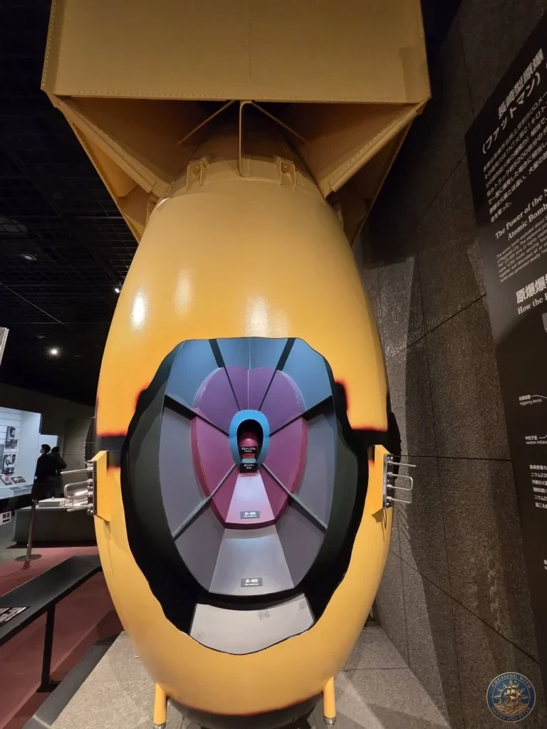 Large replica of the “Fat Man” atomic bomb displayed at the Nagasaki Atomic Bomb Museum. The yellow bomb casing is shown with a cutaway view revealing the internal plutonium core and complex detonation mechanism. Informational panels are visible beside the exhibit. Featured in Nagasaki Japan cruise port guide, things to do in Nagasaki from cruise ship, and one day in Nagasaki Japan itinerary from the Westerdam. by Danny and Peter.