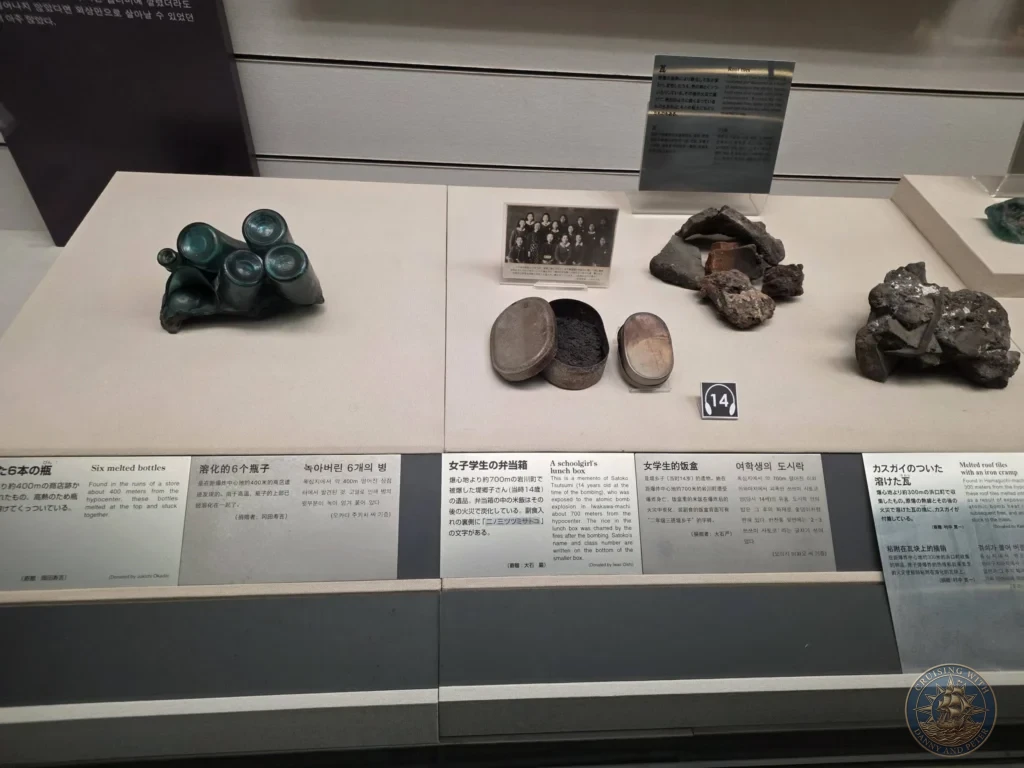 Exhibit case at the Nagasaki Atomic Bomb Museum displaying fused melted glass bottles, a deformed schoolgirl’s lunch box, melted roof tiles with iron cramps, and other heat-damaged objects from the atomic bombing. Informational panels in multiple languages explain each item. Featured in Nagasaki Japan cruise port guide, things to do in Nagasaki from cruise ship, and one day in Nagasaki Japan itinerary from the Westerdam. by Danny and Peter.