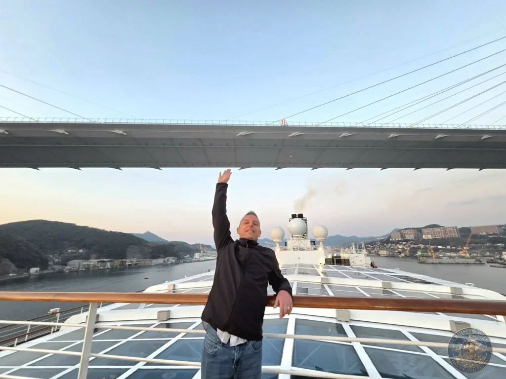 Peter waving from the deck of the Westerdam cruise ship as it sails under the Megami Bridge during sailaway from Nagasaki, Japan. The iconic bridge spans above the ship with the harbor and city in the background. Featured in Nagasaki Japan cruise port guide, things to do in Nagasaki from cruise ship, and one day in Nagasaki Japan itinerary from the Westerdam. by Danny and Peter.