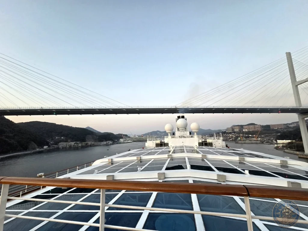 View from the deck of the Westerdam cruise ship passing directly under the Megami Bridge during sailaway from Nagasaki, Japan. The massive bridge structure spans overhead with the harbor, hills, and city visible in the background. Featured in Nagasaki Japan cruise port guide, things to do in Nagasaki from cruise ship, and one day in Nagasaki Japan itinerary from the Westerdam. by Danny and Peter.