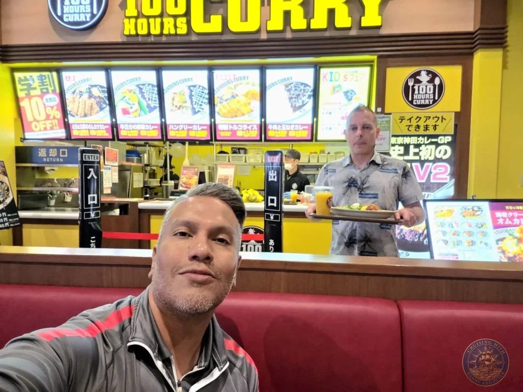 Selfie at 100 Hours Curry restaurant in Nagasaki, Japan, with Danny in the foreground and Peter holding a tray of curry in the background. The bright yellow interior features menu boards showing various curry dishes. Featured in Nagasaki Japan cruise port guide, things to do in Nagasaki from cruise ship, and one day in Nagasaki Japan itinerary from the Westerdam. by Danny and Peter.