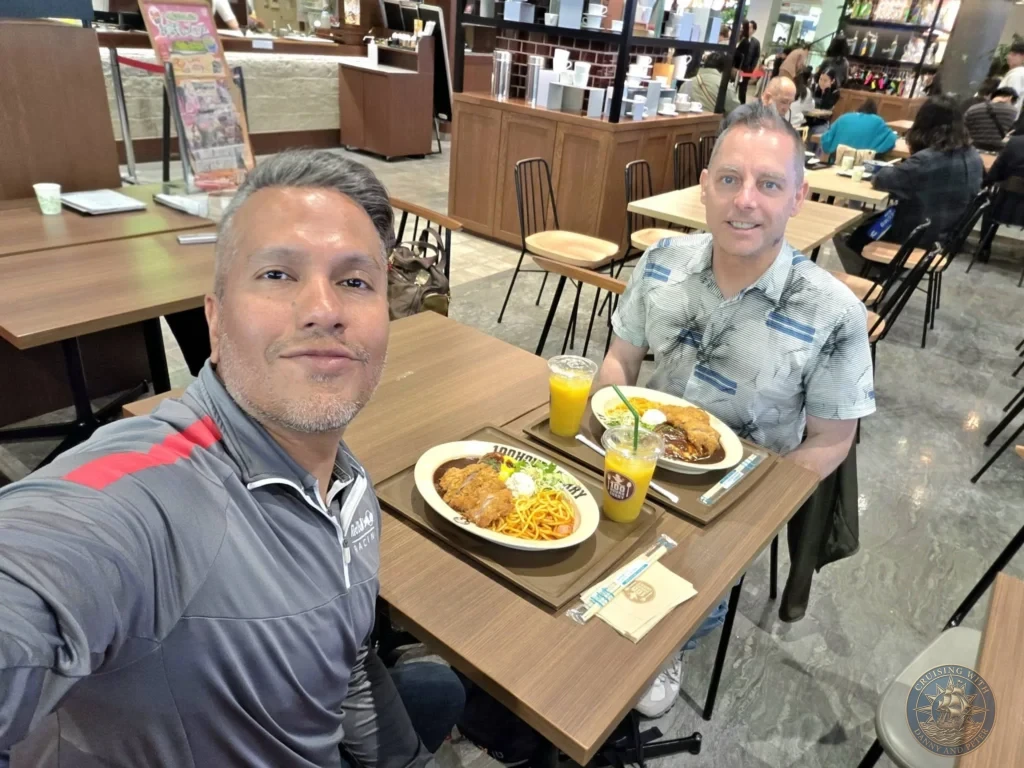 Danny and Peter enjoying a meal at 100 Hours Curry restaurant in Nagasaki, Japan. The table features plates of Japanese curry with fried pork cutlet and noodles, plus glasses of orange juice. The casual restaurant interior is visible in the background. Featured in Nagasaki Japan cruise port guide, things to do in Nagasaki from cruise ship, and one day in Nagasaki Japan itinerary from the Westerdam. by Danny and Peter.