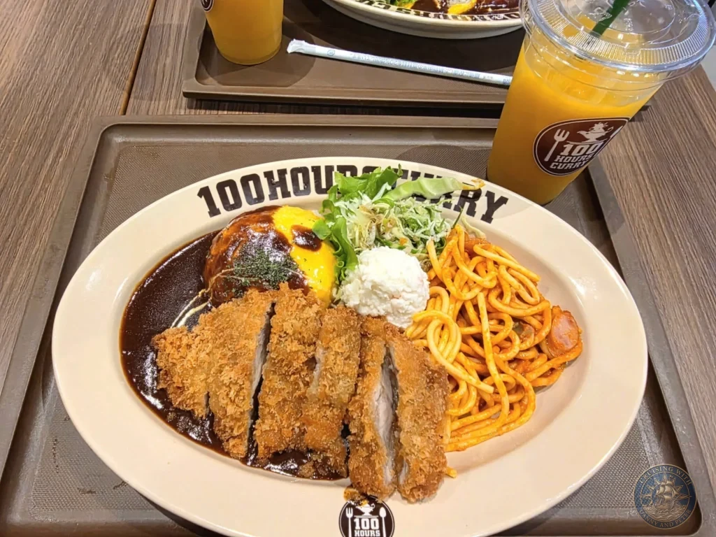 Close-up of a hearty plate of Japanese curry at 100 Hours Curry in Nagasaki, Japan — featuring crispy tonkatsu (pork cutlet), spaghetti, a soft-boiled egg, shredded cabbage, and rich brown curry sauce, served with a side of orange juice. Featured in Nagasaki Japan cruise port guide, things to do in Nagasaki from cruise ship, and one day in Nagasaki Japan itinerary from the Westerdam. by Danny and Peter.
