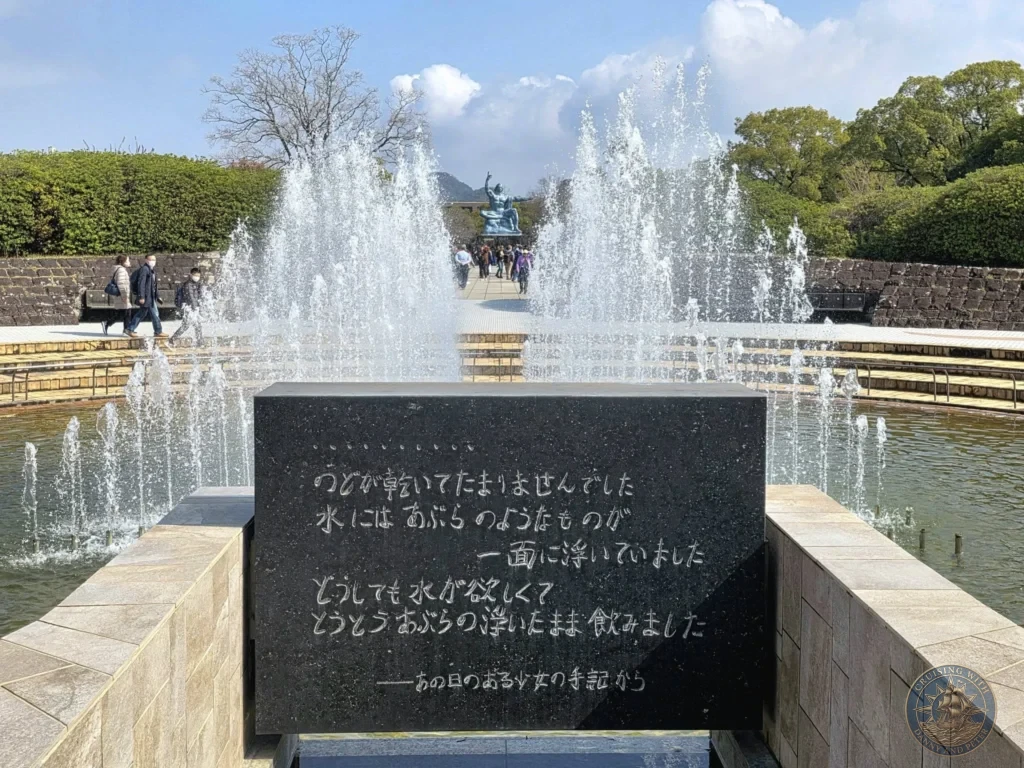 Peace Fountain with powerful water jets at Nagasaki Peace Park, Japan. In the background stands the famous Peace Statue with arms raised, surrounded by visitors on a sunny day. A black stone plaque with Japanese inscription is in the foreground. Featured in Nagasaki Japan cruise port guide, things to do in Nagasaki from cruise ship, and one day in Nagasaki Japan itinerary from the Westerdam. by Danny and Peter.