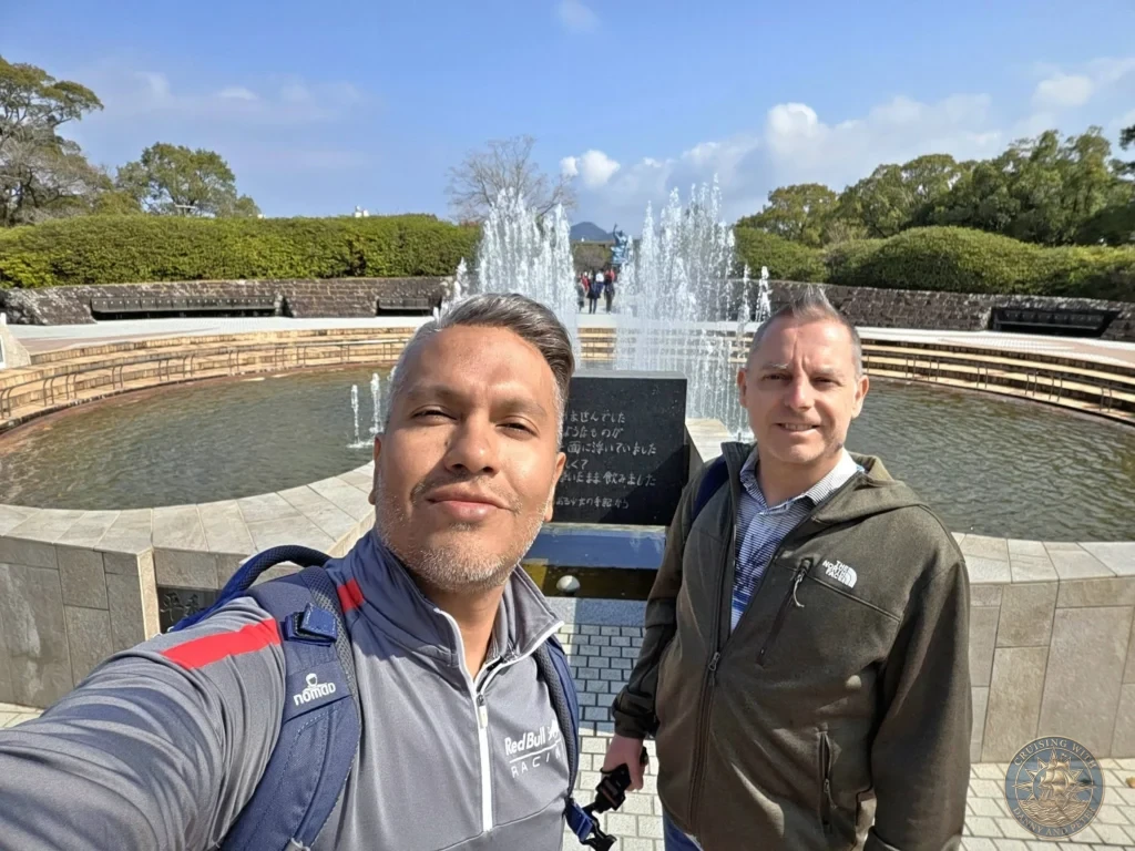 Selfie of Danny and Peter at the Peace Fountain in Nagasaki Peace Park, Japan. The fountain jets are spraying water behind them, with the famous Peace Statue visible in the distance. Featured in Nagasaki Japan cruise port guide, things to do in Nagasaki from cruise ship, and one day in Nagasaki Japan itinerary from the Westerdam. by Danny and Peter.