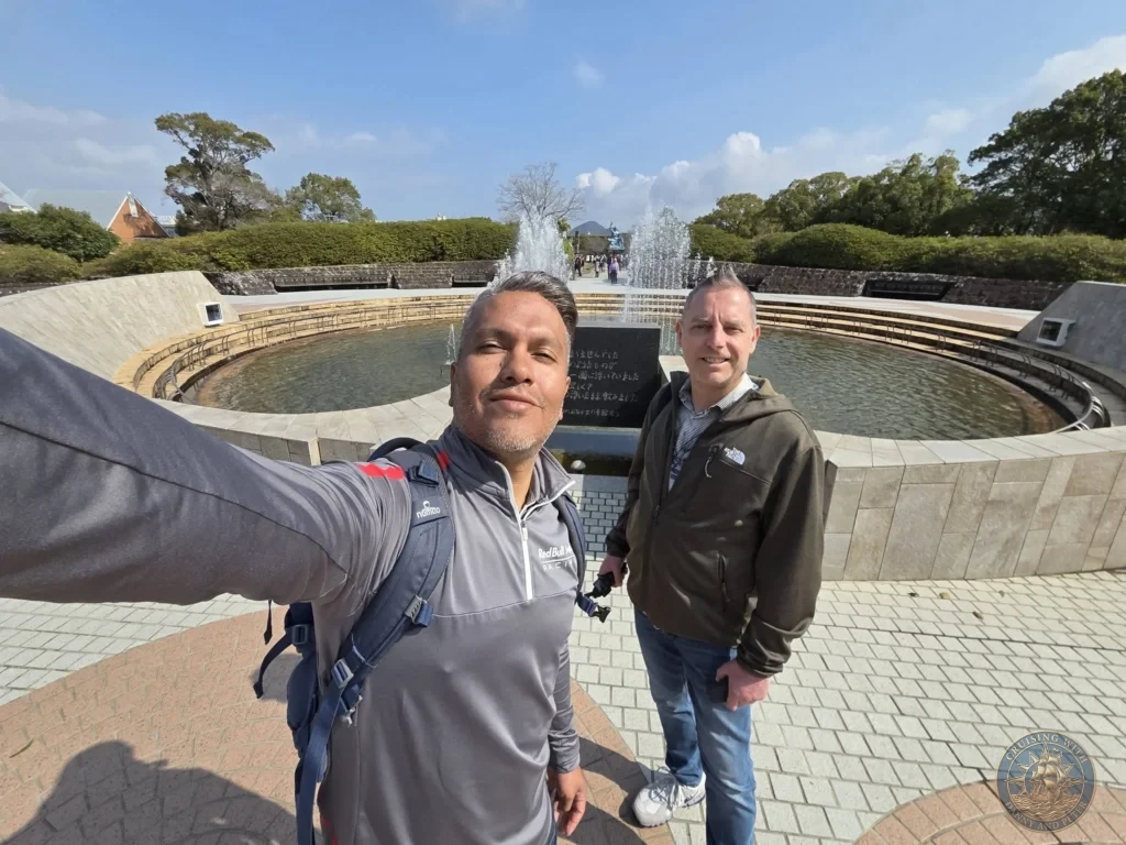 Selfie of Danny and Peter at the Peace Fountain in Nagasaki Peace Park, Japan. The circular fountain with spraying water jets and the iconic Peace Statue are visible in the background under a clear blue sky. Featured in Nagasaki Japan cruise port guide, things to do in Nagasaki from cruise ship, and one day in Nagasaki Japan itinerary from the Westerdam. by Danny and Peter.