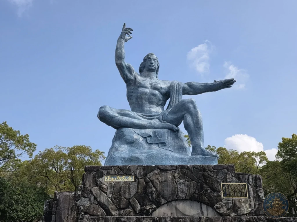 The iconic Peace Statue at Nagasaki Peace Park, Japan — a large bronze figure with one arm raised to the sky and the other extended outward, seated on a stone base. Featured in Nagasaki Japan cruise port guide, things to do in Nagasaki from cruise ship, and one day in Nagasaki Japan itinerary from the Westerdam. by Danny and Peter.