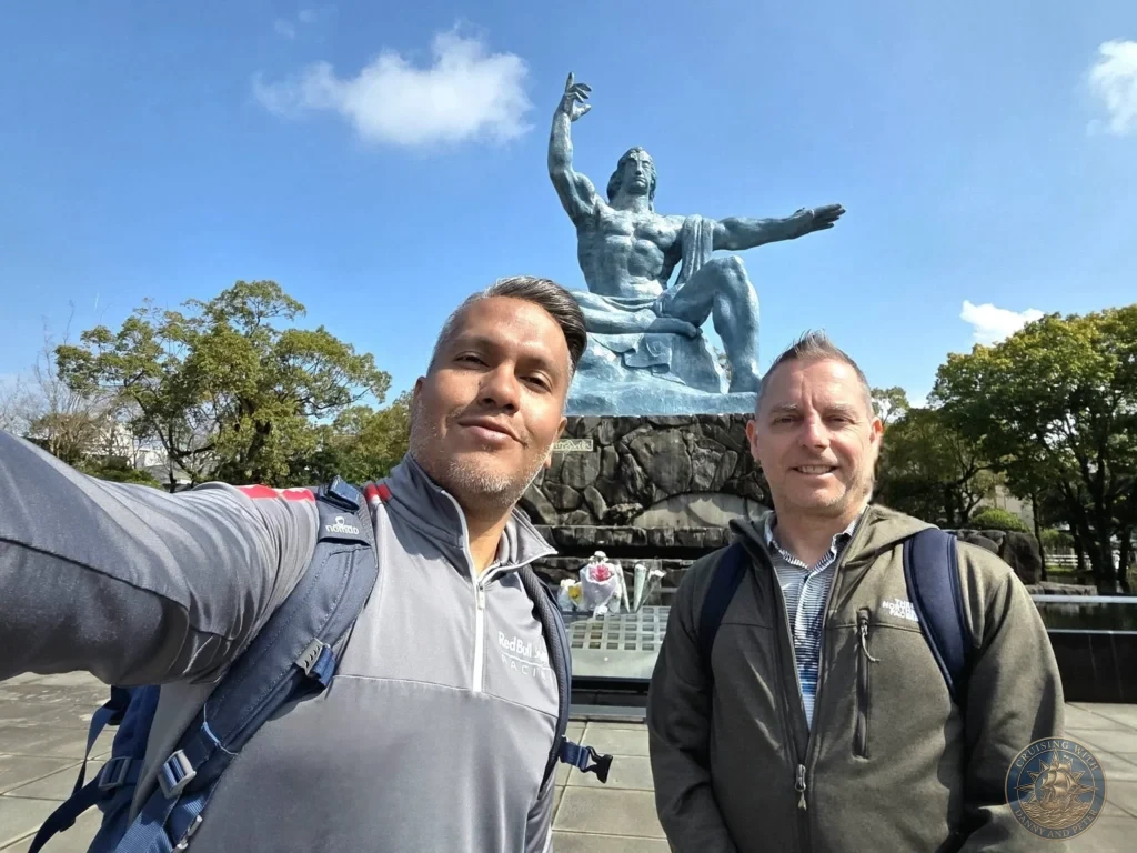 Selfie of Danny and Peter in front of the iconic Peace Statue at Nagasaki Peace Park, Japan. The large bronze statue with one arm raised toward the sky stands prominently in the background under a clear blue sky. Featured in Nagasaki Japan cruise port guide, things to do in Nagasaki from cruise ship, and one day in Nagasaki Japan itinerary from the Westerdam. by Danny and Peter.