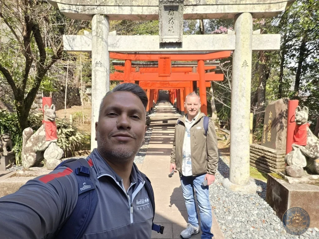 Selfie of Danny and Peter in front of the iconic red torii gates at Suwa Shrine in Nagasaki, Japan. A long tunnel of bright orange torii leads up the stairs, flanked by stone fox statues. Featured in Nagasaki Japan cruise port guide, things to do in Nagasaki from cruise ship, and one day in Nagasaki Japan itinerary from the Westerdam. by Danny and Peter.