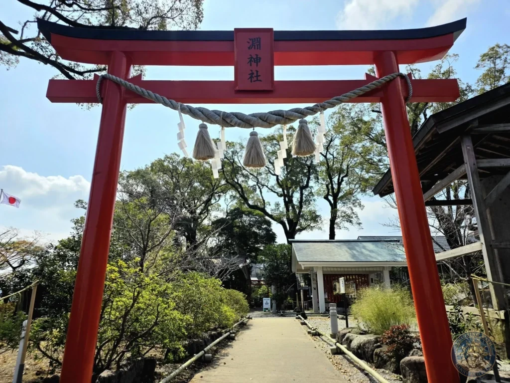 Large red torii gate with shimenawa rope and white shide streamers at Suwa Shrine in Nagasaki, Japan. A paved path leads through the gate toward the shrine buildings, surrounded by trees under a clear blue sky. Featured in Nagasaki Japan cruise port guide, things to do in Nagasaki from cruise ship, and one day in Nagasaki Japan itinerary from the Westerdam. by Danny and Peter.