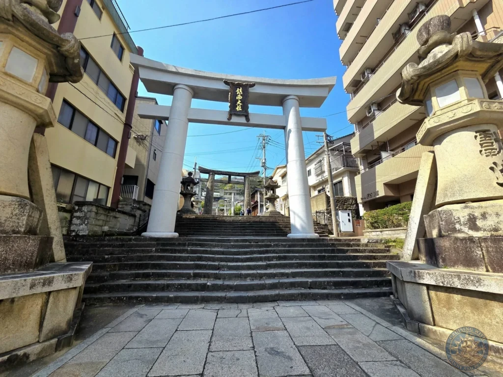 Large white torii gate with a black sign at Suwa Shrine in Nagasaki, Japan. Wide stone stairs lead upward through the gate, flanked by traditional stone lanterns. Urban buildings rise on both sides under a clear blue sky. Featured in Nagasaki Japan cruise port guide, things to do in Nagasaki from cruise ship, and one day in Nagasaki Japan itinerary from the Westerdam. by Danny and Peter.
