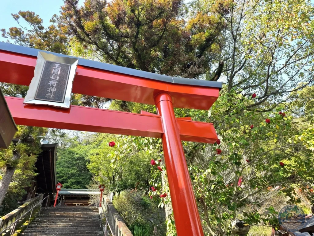Bright red torii gate with a sign reading “Suwa Shrine” at Suwa Shrine in Nagasaki, Japan. Stone stairs lead upward through the gate, surrounded by lush green trees and foliage under a clear blue sky. Featured in Nagasaki Japan cruise port guide, things to do in Nagasaki from cruise ship, and one day in Nagasaki Japan itinerary from the Westerdam. by Danny and Peter.