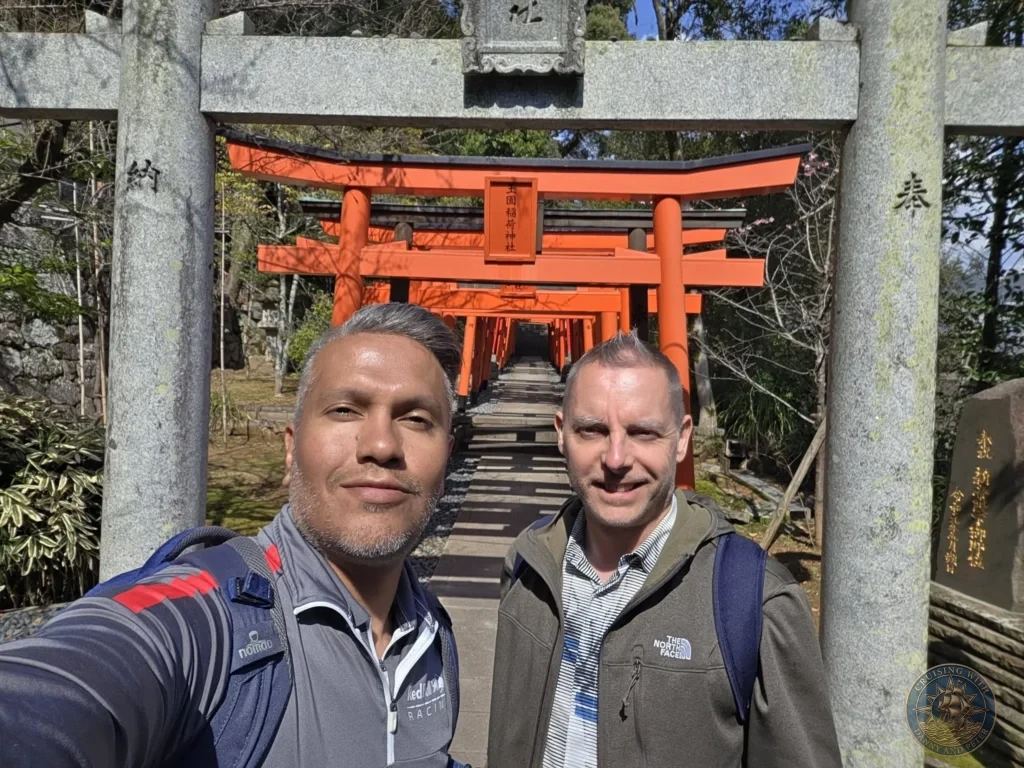 Selfie of Danny and Peter standing in front of the large stone torii gate at Suwa Shrine in Nagasaki, Japan, with the long tunnel of red torii gates stretching into the background. Featured in Nagasaki Japan cruise port guide, things to do in Nagasaki from cruise ship, and one day in Nagasaki Japan itinerary from the Westerdam. by Danny and Peter.