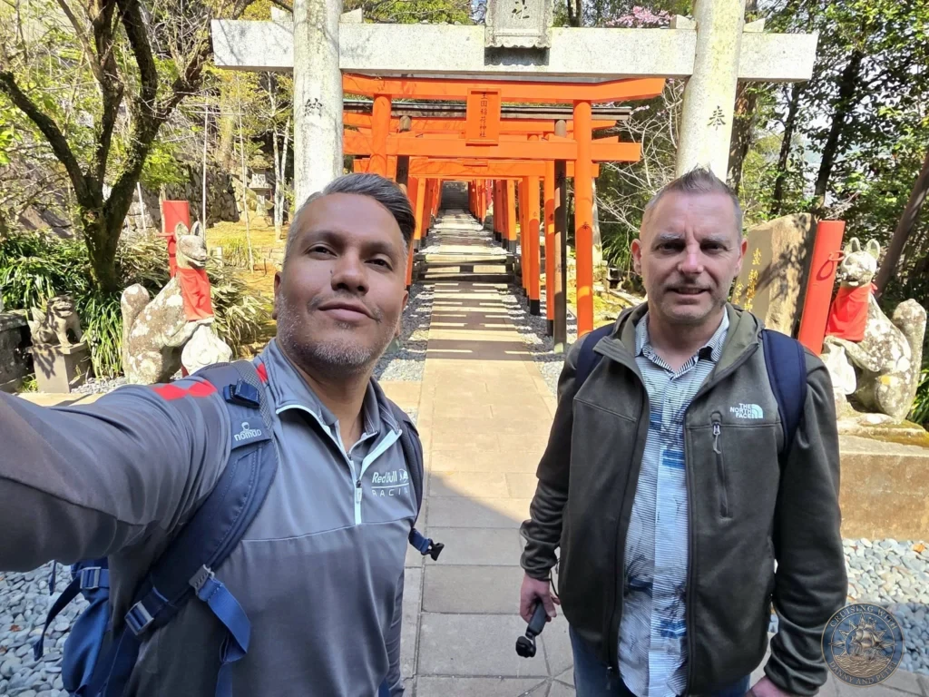 Selfie of Danny and Peter at the entrance to Suwa Shrine in Nagasaki, Japan, standing in front of the large stone torii gate with the long tunnel of vibrant red torii gates stretching into the background. Featured in Nagasaki Japan cruise port guide, things to do in Nagasaki from cruise ship, and one day in Nagasaki Japan itinerary from the Westerdam. by Danny and Peter.