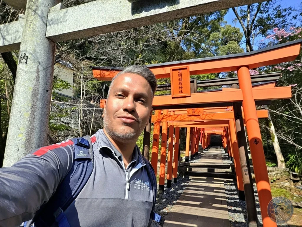 Selfie of Danny inside the long tunnel of red torii gates at Suwa Shrine in Nagasaki, Japan. The vibrant orange gates line the path uphill, surrounded by trees under a bright sky. Featured in Nagasaki Japan cruise port guide, things to do in Nagasaki from cruise ship, and one day in Nagasaki Japan itinerary from the Westerdam. by Danny and Peter.