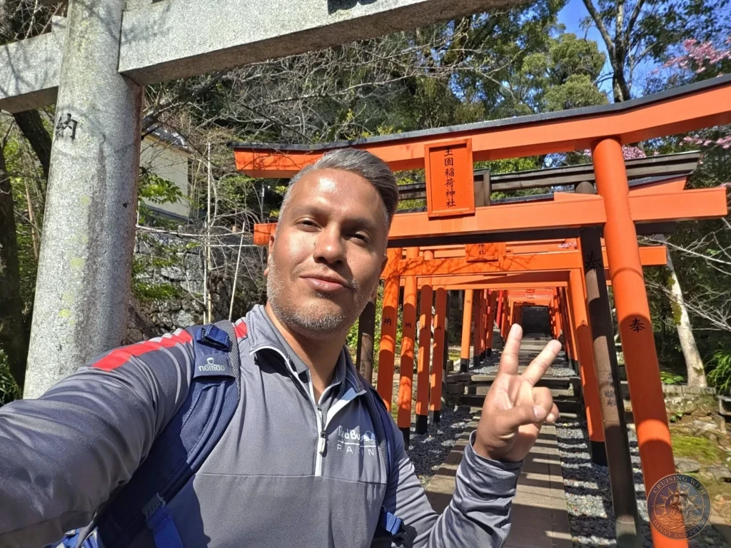 Selfie of Danny making a peace sign inside the long tunnel of red torii gates at Suwa Shrine in Nagasaki, Japan. The vibrant orange gates line the path uphill under a bright blue sky. Featured in Nagasaki Japan cruise port guide, things to do in Nagasaki from cruise ship, and one day in Nagasaki Japan itinerary from the Westerdam. by Danny and Peter.