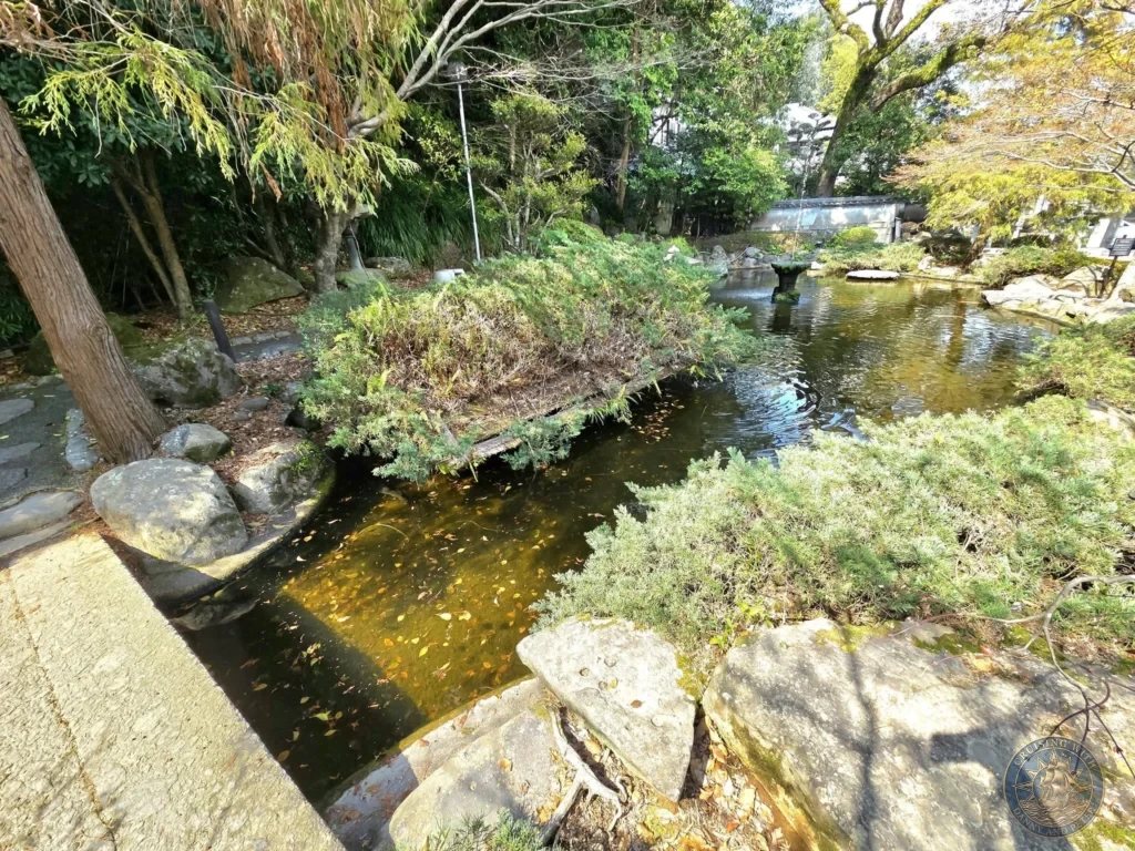 Serene garden pond with a stone fountain at Suwa Shrine in Nagasaki, Japan. The pond is surrounded by rocks, lush greenery, and trees, creating a peaceful traditional Japanese garden scene. Featured in Nagasaki Japan cruise port guide, things to do in Nagasaki from cruise ship, and one day in Nagasaki Japan itinerary from the Westerdam. by Danny and Peter.