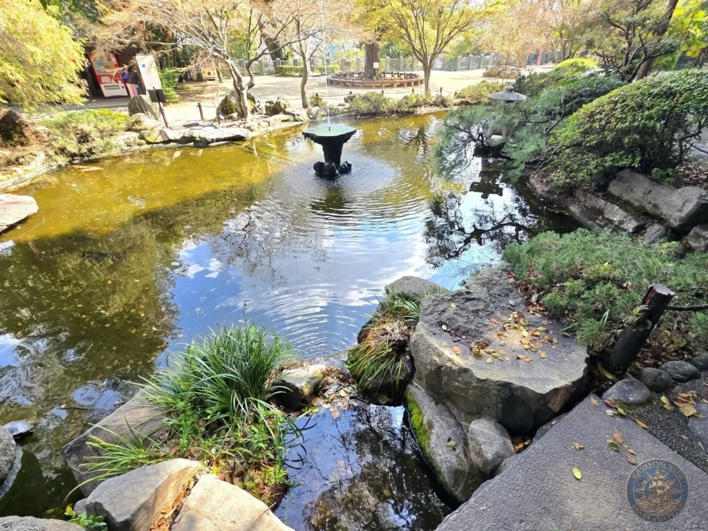 Serene pond with a central stone fountain at Suwa Shrine in Nagasaki, Japan. The pond is surrounded by large rocks, green plants, and autumn-colored trees, with reflections of the sky and foliage on the water. Featured in Nagasaki Japan cruise port guide, things to do in Nagasaki from cruise ship, and one day in Nagasaki Japan itinerary from the Westerdam. by Danny and Peter.
