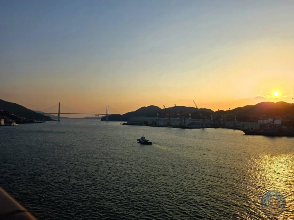 Stunning sunset over Nagasaki harbor viewed from the Westerdam cruise ship. The sun sets behind the hills with the Megami Bridge visible in the distance, a small tugboat on the water, and golden reflections on the sea. Featured in Nagasaki Japan cruise port guide, Westerdam Nagasaki docking location, things to do in Nagasaki from cruise ship, and one day in Nagasaki Japan itinerary. by Danny and Peter.