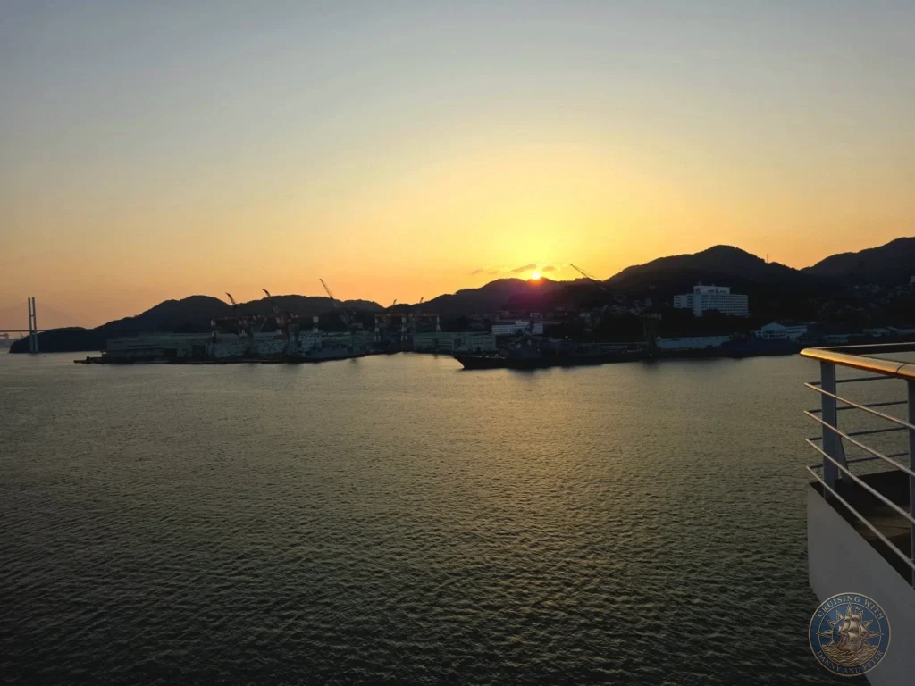 Beautiful sunset viewed from the deck of the Westerdam in Nagasaki harbor, Japan. The sun sets behind the hills with golden light reflecting on the water, industrial cranes and port facilities silhouetted in the foreground, and the Megami Bridge visible in the distance. Featured in Nagasaki Japan cruise port guide, Westerdam Nagasaki docking location, things to do in Nagasaki from cruise ship, and one day in Nagasaki Japan itinerary. by Danny and Peter.