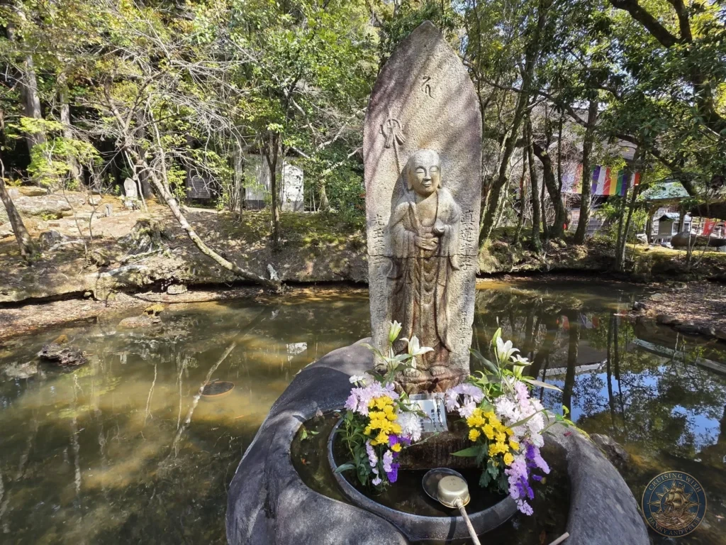 Stone Jizo Bodhisattva statue with vibrant flower offerings placed in a pond at Chikurin Temple in Kochi, Japan. Peaceful temple memorial feature surrounded by forest. Kochi Cruise Port Guide | Things to do in Kochi from cruise ship | One day in Kochi Japan itinerary by Danny and Peter.