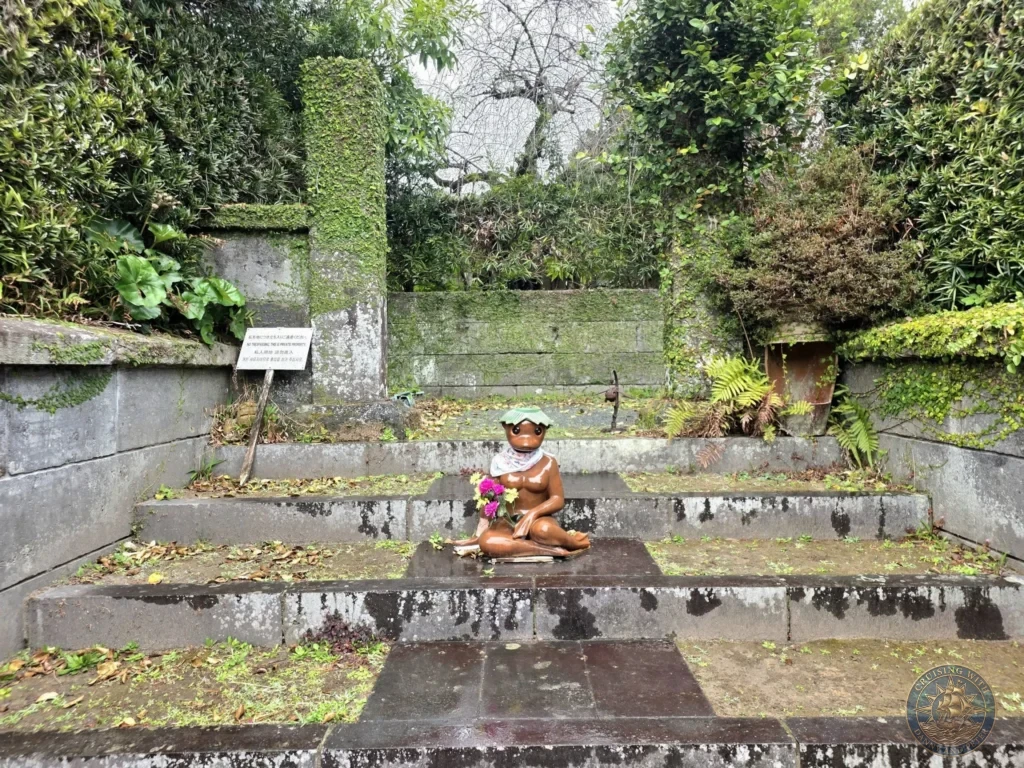 Charming kappa (water imp) statue wearing a green hat and scarf, seated on stone steps surrounded by mossy walls and lush greenery in the historic Chiran Samurai District. Quirky cultural highlight during a Kagoshima shore excursion. Kagoshima Cruise Port Guide | Things to do in Kagoshima from cruise ship | One day in Kagoshima Japan itinerary by Danny and Peter.