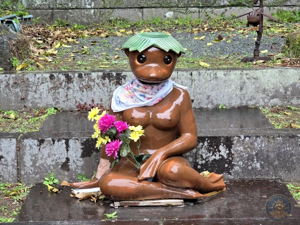 Close-up of the charming kappa statue with green hat and floral scarf, seated on stone steps with fresh flowers in the historic Chiran Samurai District. Popular quirky sight for visitors exploring the samurai gardens during a Kagoshima cruise stop. Kagoshima Cruise Port Guide | Things to do in Kagoshima from cruise ship | One day in Kagoshima Japan itinerary by Danny and Peter.