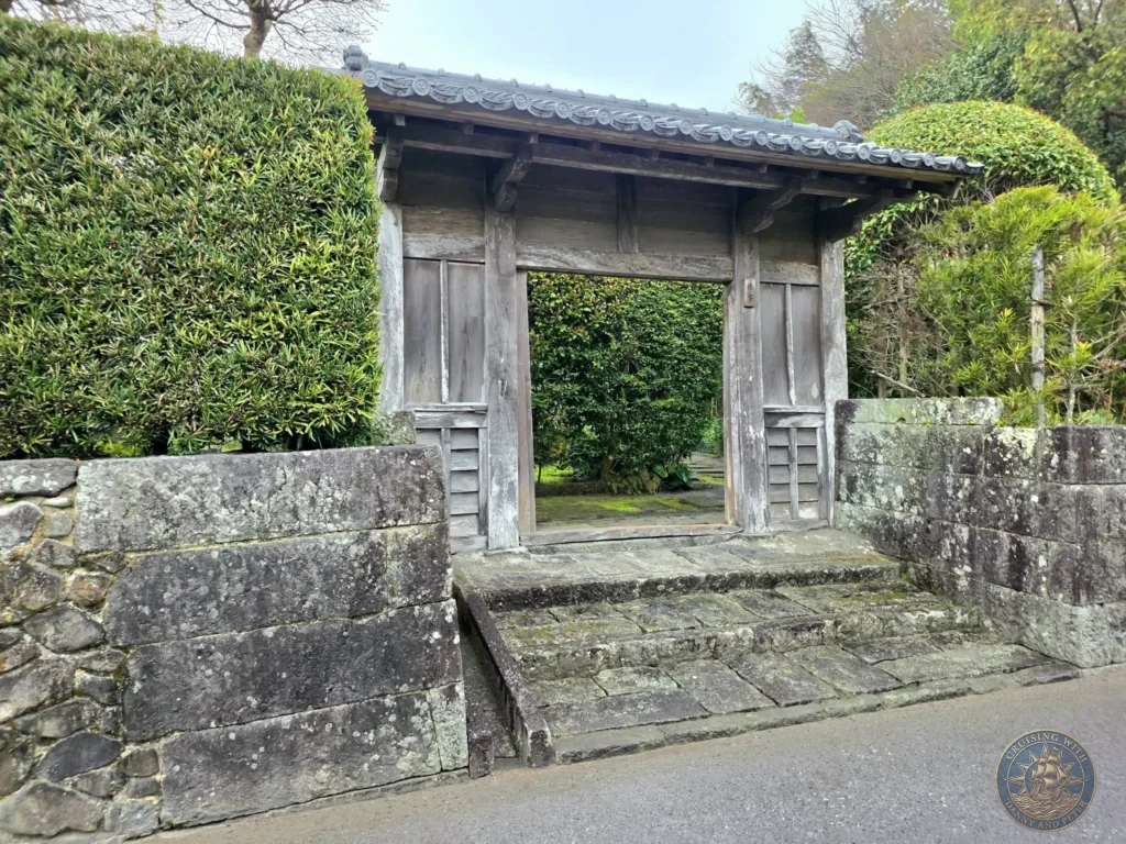 Traditional wooden entrance gate with tiled roof leading into a samurai residence garden, framed by stone walls and tall manicured hedges in the historic Chiran Samurai District. Iconic view of preserved Edo-period architecture in Kagoshima, Japan. Kagoshima Cruise Port Guide | Things to do in Kagoshima from cruise ship | One day in Kagoshima Japan itinerary by Danny and Peter.