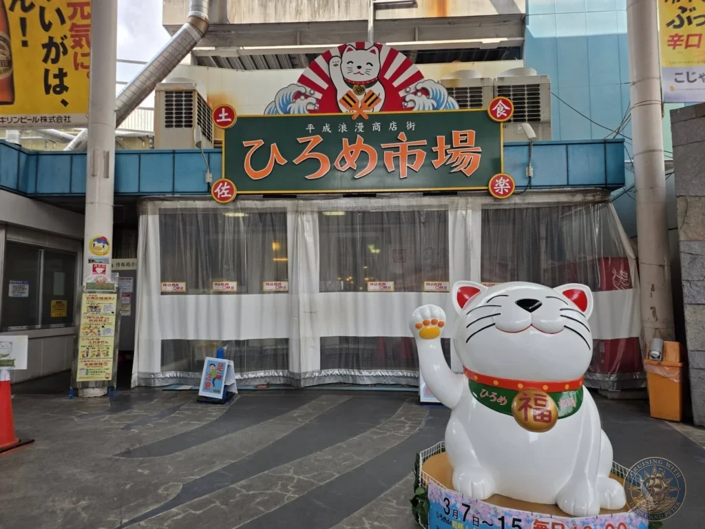 Entrance to Hirome Market in Kochi, Japan, with large Maneki-neko lucky cat statue and vibrant signage. Iconic local food market. Kochi Cruise Port Guide | Things to do in Kochi from cruise ship | One day in Kochi Japan itinerary by Danny and Peter.