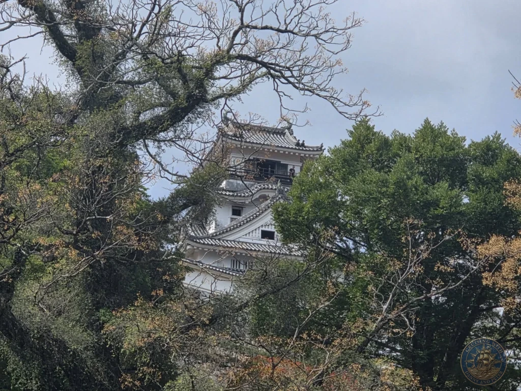 Historic Kochi Castle keep framed by tree branches in Kochi, Japan. Scenic view of the white castle surrounded by lush greenery. Kochi Cruise Port Guide | Things to do in Kochi from cruise ship | One day in Kochi Japan itinerary by Danny and Peter.