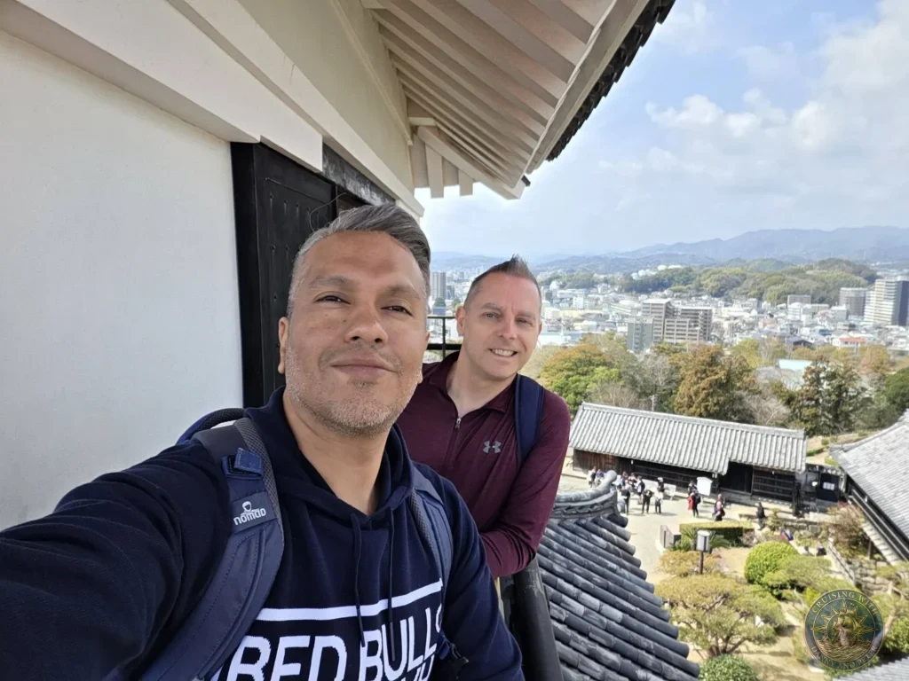 Danny and Peter taking a selfie on the balcony of Kochi Castle with sweeping city and mountain views in Kochi, Japan. Iconic historic viewpoint. Kochi Cruise Port Guide | Things to do in Kochi from cruise ship | One day in Kochi Japan itinerary by Danny and Peter.