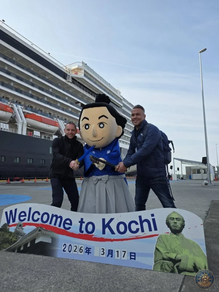 Danny and Peter posing with local mascot in front of Holland America Westerdam docked at Kochi Port, Japan. Cruise ship arrival with welcome sign. Kochi Cruise Port Guide | Things to do in Kochi from cruise ship | One day in Kochi Japan itinerary by Danny and Peter.