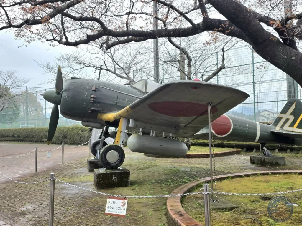 Historic kamikaze aircraft on outdoor display at the Chiran Peace Museum for Kamikaze Pilots, with cherry blossom trees and memorial statue nearby. Powerful exhibit in the Chiran Samurai District, Kagoshima, Japan. Kagoshima Cruise Port Guide | Things to do in Kagoshima from cruise ship | One day in Kagoshima Japan itinerary by Danny and Peter.
