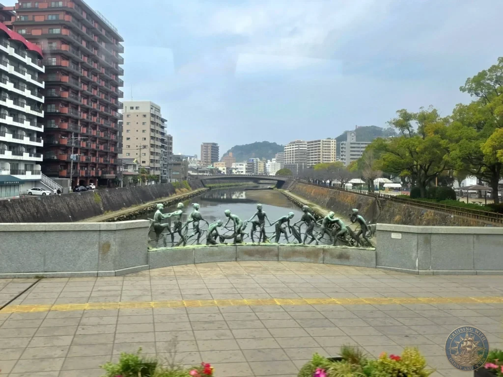 Bronze sculpture of figures pulling a net along the Kotsuki River promenade in central Kagoshima City, with modern buildings and a bridge in the background. Popular riverside attraction and photo spot near the city center. Kagoshima Cruise Port Guide | Things to do in Kagoshima from cruise ship | One day in Kagoshima Japan itinerary by Danny and Peter.