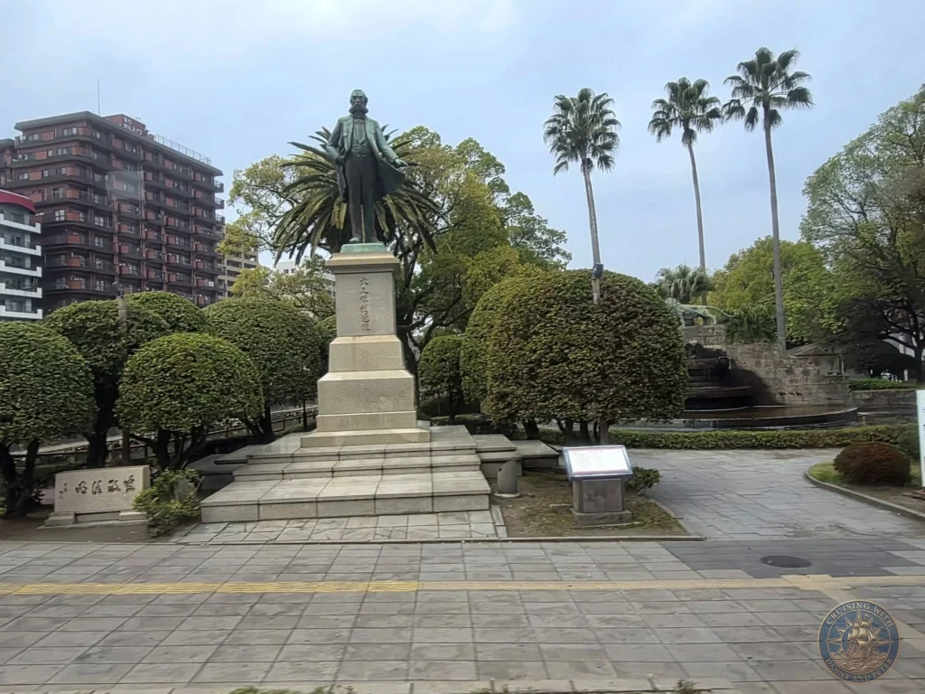 Iconic bronze statue of Saigō Takamori, the legendary "Last Samurai" and Satsuma leader, standing prominently in a park in central Kagoshima City with palm trees and modern buildings in the background. Major historical landmark and popular sightseeing spot. Kagoshima Cruise Port Guide | Things to do in Kagoshima from cruise ship | One day in Kagoshima Japan itinerary by Danny and Peter.