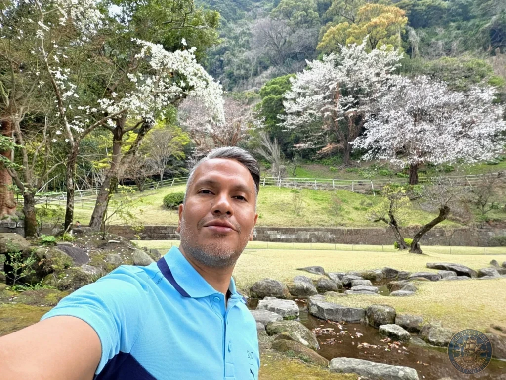 Danny taking a selfie at Sengan-en (Iso Garden) with blooming cherry blossoms, traditional landscaping, and lush green hills in the background. Beautiful spring scenery at the historic UNESCO World Heritage samurai garden in Kagoshima, Japan. Kagoshima Cruise Port Guide | Things to do in Kagoshima from cruise ship | One day in Kagoshima Japan itinerary by Danny and Peter.