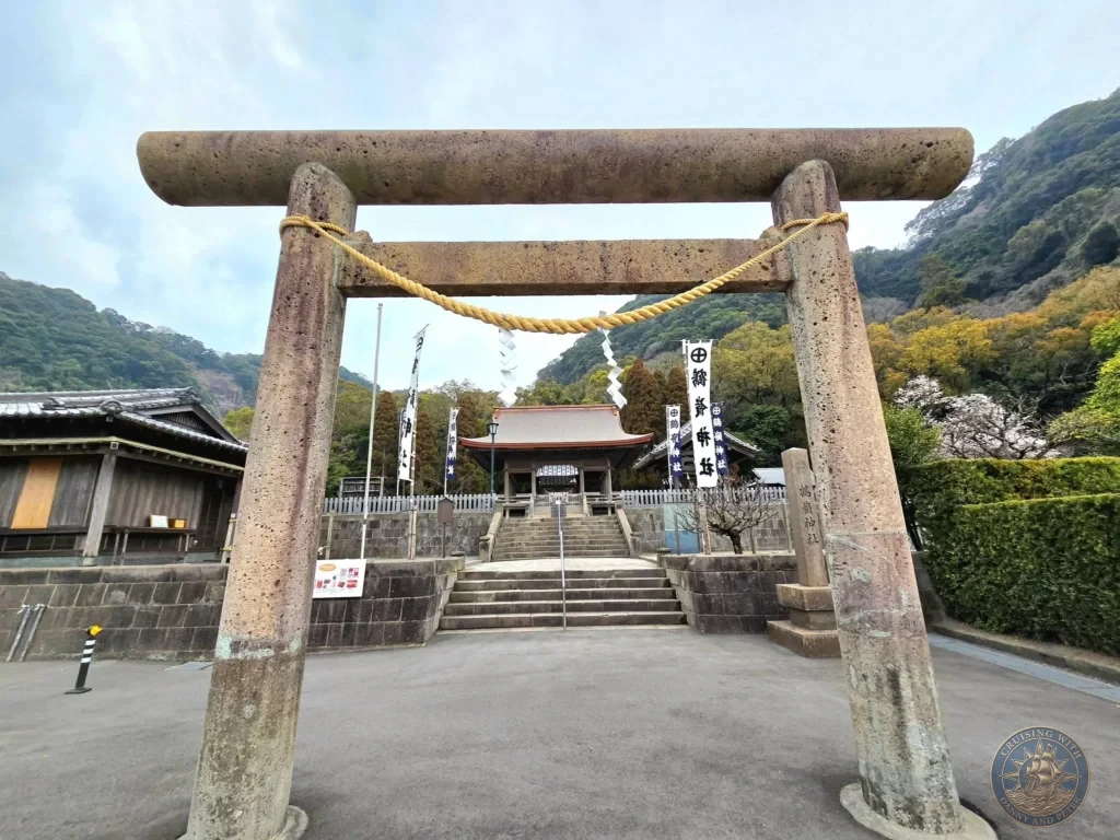 Large stone torii gate with shimenawa rope marking the entrance to Tsurugane Shrine at Sengan-en (Iso Garden), the historic UNESCO World Heritage samurai villa in Kagoshima, Japan. Scenic shrine approach with forested hills in the background. Kagoshima Cruise Port Guide | Things to do in Kagoshima from cruise ship | One day in Kagoshima Japan itinerary by Danny and Peter.
