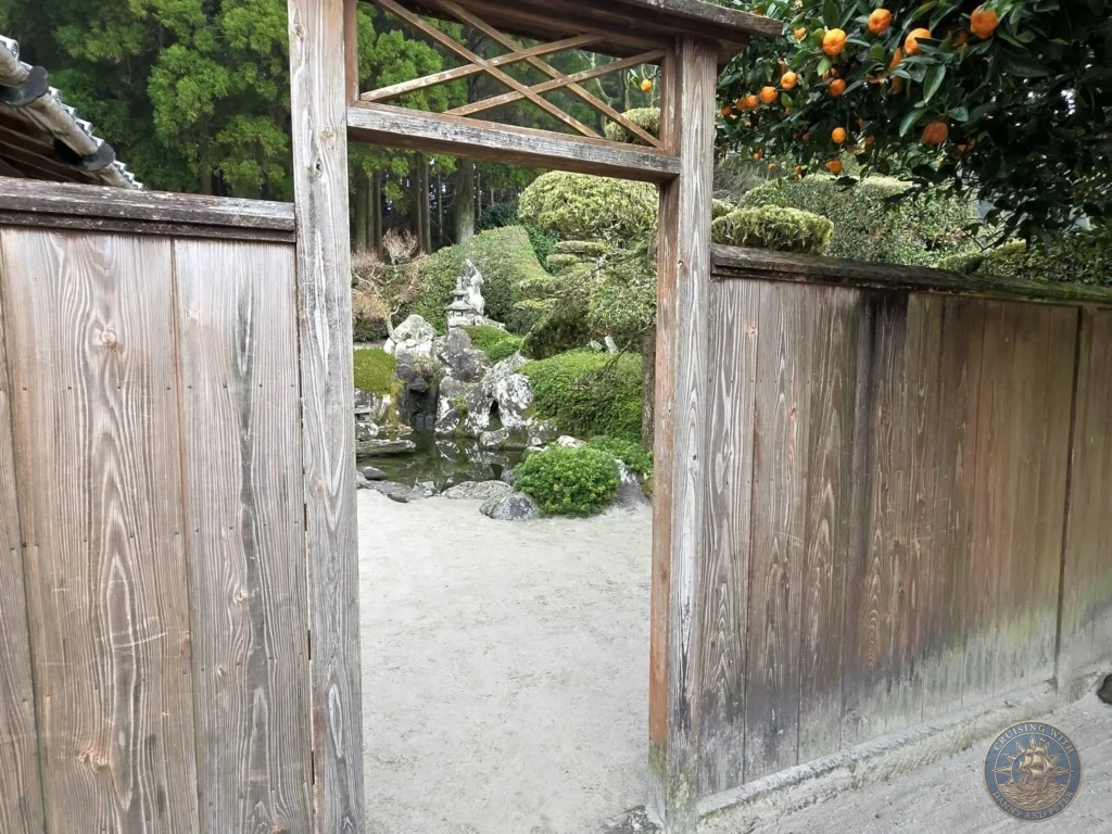 Traditional wooden gate framing the view into Tsukiyama-sensui Garden 7 (Pond & Waterfall) at Chiran Samurai District in Kagoshima, Japan. Serene pond, rocks, stone lantern, and citrus trees with ripe fruit. Kagoshima Cruise Port Guide | Things to do in Kagoshima from cruise ship | One day in Kagoshima Japan itinerary by Danny and Peter.