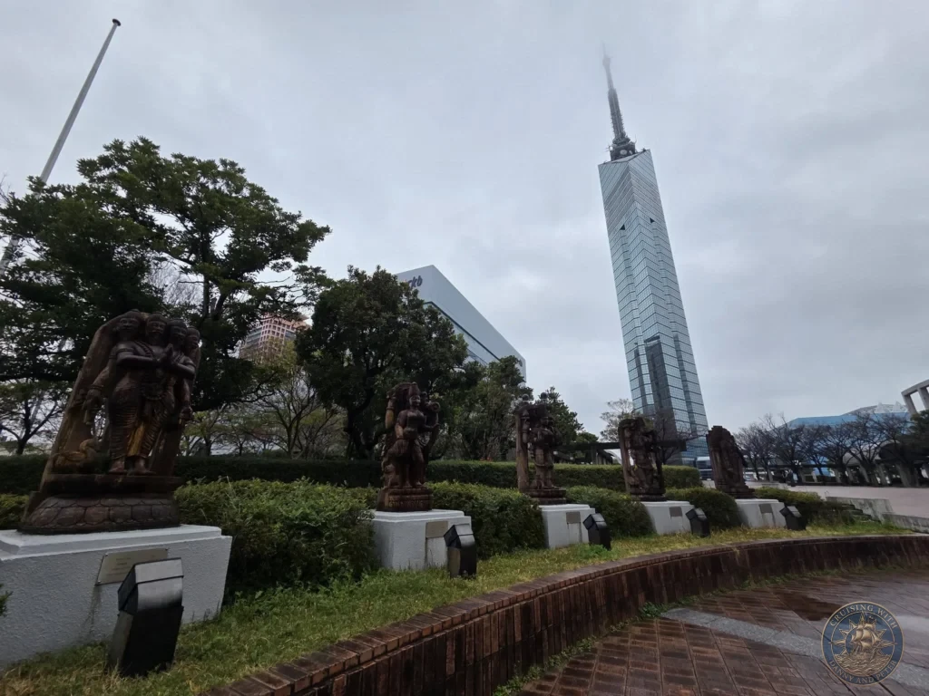 Fukuoka Tower soaring into the cloudy sky with sculptures in the foreground park, Fukuoka, Japan. City landmark. Fukuoka Cruise Port Guide | Things to do in Fukuoka from cruise ship | One day in Fukuoka Japan itinerary by Danny and Peter.