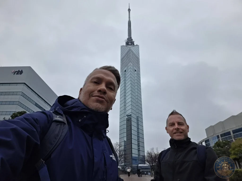 Danny and Peter taking a selfie with the tall Fukuoka Tower in the background on a cloudy day in Fukuoka, Japan. City landmark. Fukuoka Cruise Port Guide | Things to do in Fukuoka from cruise ship | One day in Fukuoka Japan itinerary by Danny and Peter.