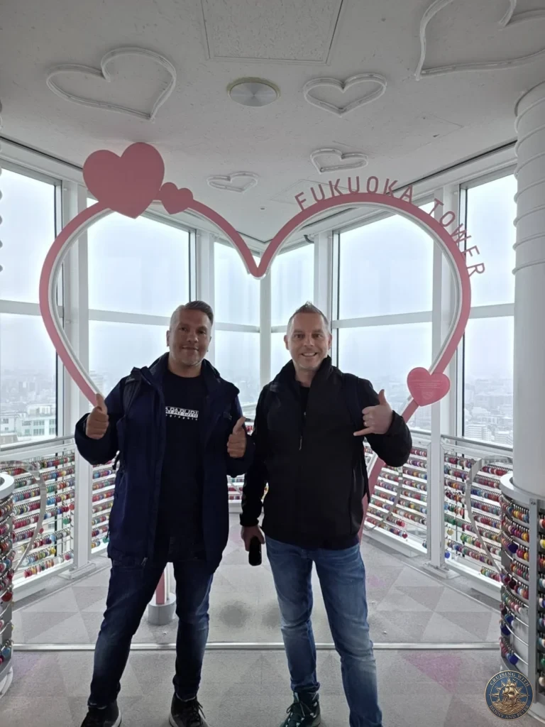 Danny and Peter smiling with thumbs up in front of the heart-shaped frame at Fukuoka Tower observation deck, Japan. Landmark moment. Fukuoka Cruise Port Guide | Things to do in Fukuoka from cruise ship | One day in Fukuoka Japan itinerary by Danny and Peter.