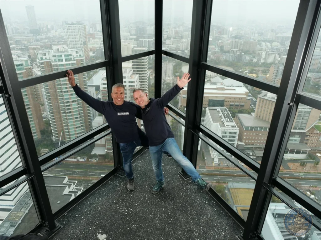 Danny and Peter posing with arms outstretched in front of the large window at Fukuoka Tower observation deck, Japan. City view moment. Fukuoka Cruise Port Guide | Things to do in Fukuoka from cruise ship | One day in Fukuoka Japan itinerary by Danny and Peter.