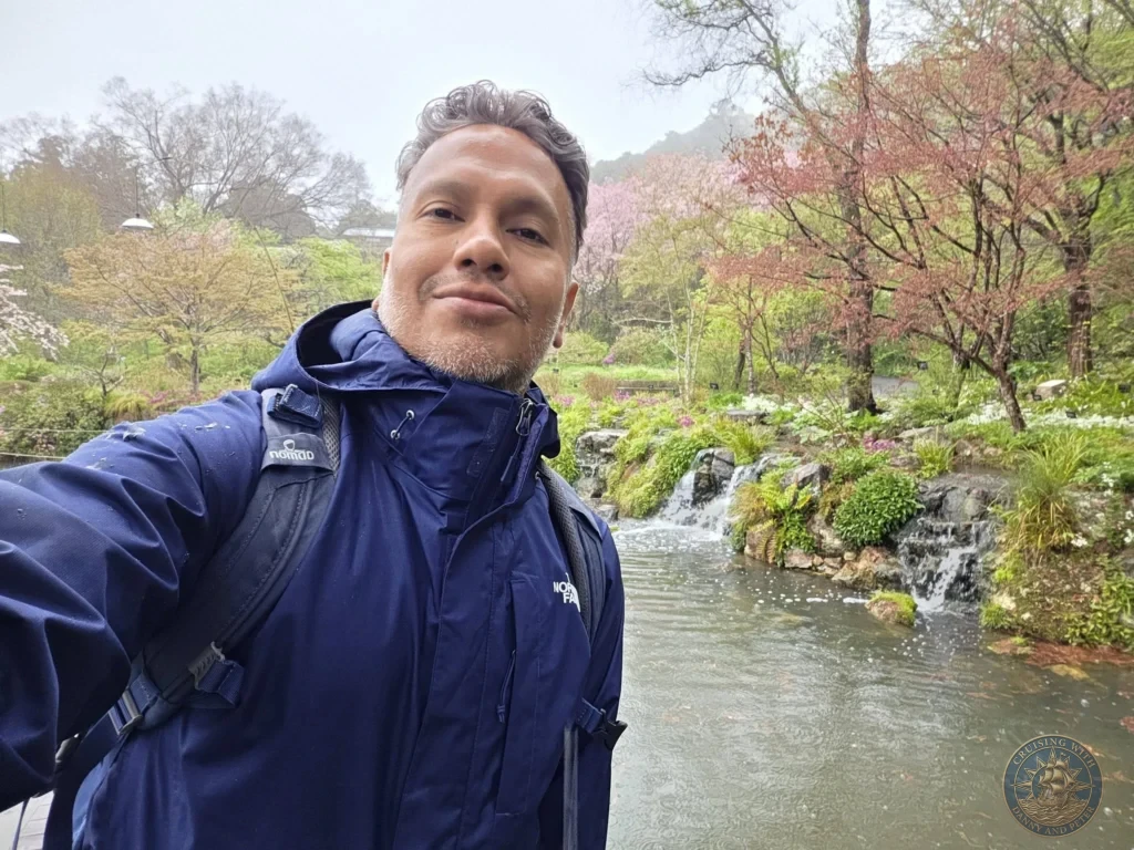 Danny taking a selfie by scenic pond with waterfalls and cherry blossoms at Makino Botanical Garden in Kochi, Japan. Spring garden view. Kochi Cruise Port Guide | Things to do in Kochi from cruise ship | One day in Kochi Japan itinerary by Danny and Peter.