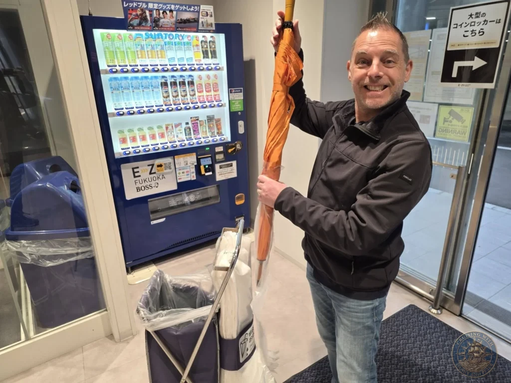 Peter smiling while holding an orange umbrella next to a blue vending machine in Fukuoka, Japan. Cruise port everyday scene. Fukuoka Cruise Port Guide | Things to do in Fukuoka from cruise ship | One day in Fukuoka Japan itinerary by Danny and Peter.