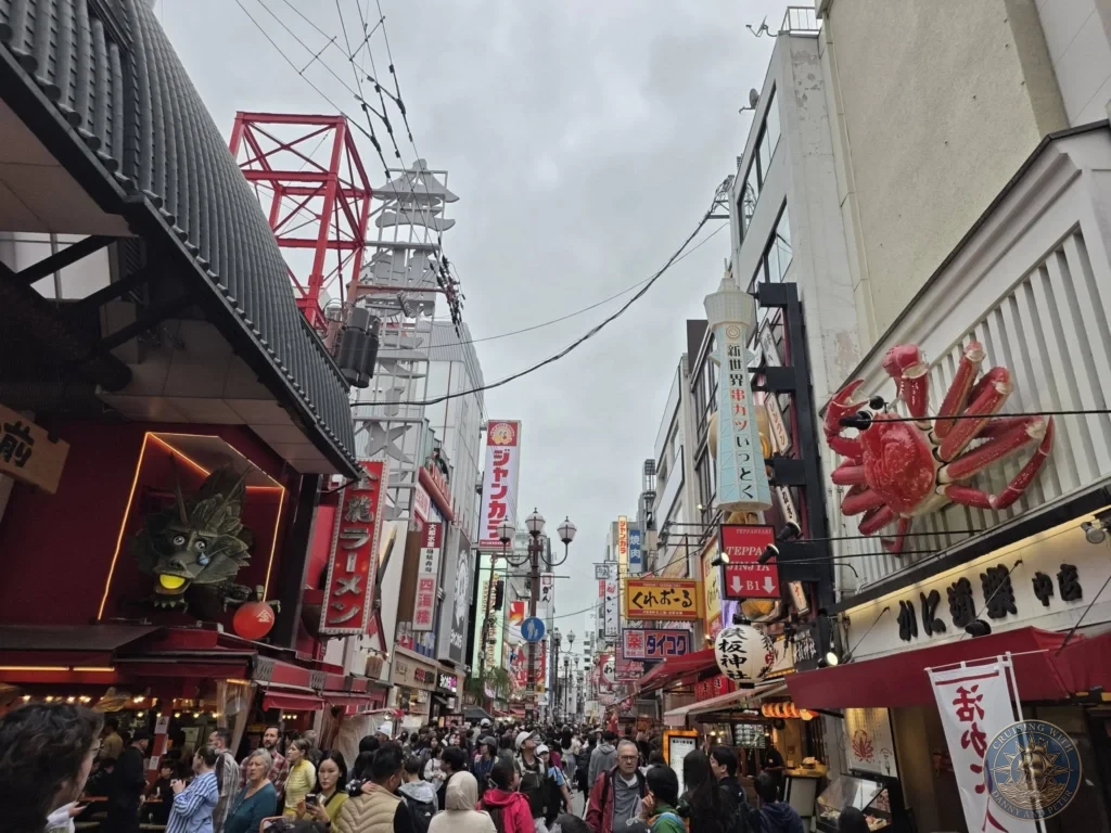 Kobe Japan Cruise Port Guide 2026 – Insider Tips & One Day Itinerary 16 Busy Dotonbori street in Osaka filled with crowds, neon signs, a giant red crab, and restaurant facades under a cloudy sky. Vibrant Osaka nightlife area from Kobe port day. Kobe Japan Cruise Port Guide | DIY Kobe Cruise Port to City Center | Things to do in Kobe from cruise ship | Westerdam Kobe Docking Location | One day in Kobe Japan itinerary by Danny and Peter.