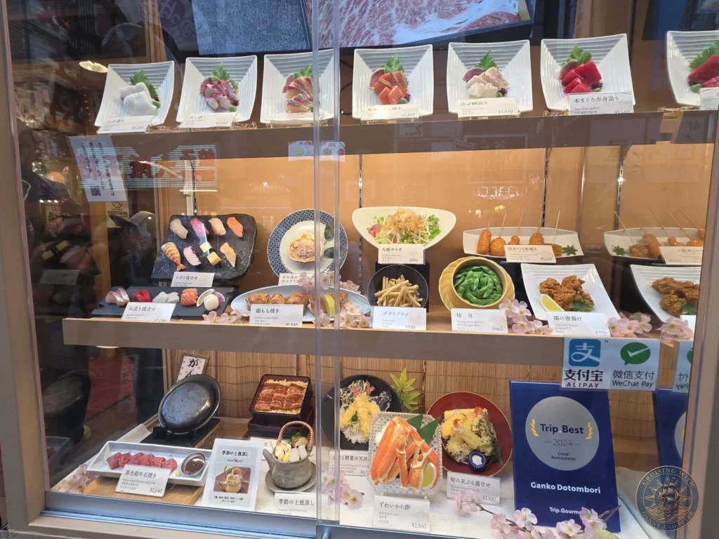 Kobe Japan Cruise Port Guide 2026 – Insider Tips & One Day Itinerary 96 Glass display window at Ganko Dotonbori showing an array of Japanese dishes including sashimi, sushi, crab, tempura, and grilled items with prices in Osaka, Japan. Restaurant showcase from Kobe port day. Kobe Japan Cruise Port Guide | DIY Kobe Cruise Port to City Center | Things to do in Kobe from cruise ship | Westerdam Kobe Docking Location | One day in Kobe Japan itinerary by Danny and Peter.
