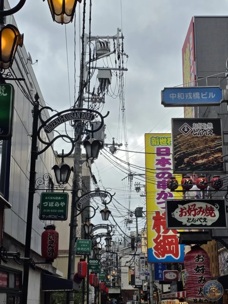 Kobe Japan Cruise Port Guide 2026 – Insider Tips & One Day Itinerary 145 Looking down Hozenji Koisan Dori in Osaka with decorative street lamps, red lanterns, restaurant signs, and overhead wires under a cloudy sky. Lively Osaka alley from Kobe port day. Kobe Japan Cruise Port Guide | DIY Kobe Cruise Port to City Center | Things to do in Kobe from cruise ship | Westerdam Kobe Docking Location | One day in Kobe Japan itinerary by Danny and Peter.