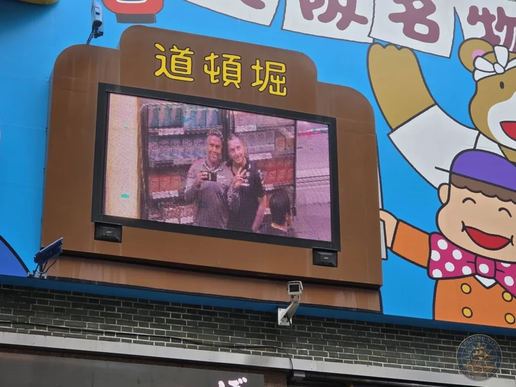 Kobe Japan Cruise Port Guide 2026 – Insider Tips & One Day Itinerary 98 Large outdoor LED screen in Dotonbori displaying a photo of Danny and Peter posing together under the Dotonbori sign in Osaka, Japan. Fun street moment from Kobe port day. Kobe Japan Cruise Port Guide | DIY Kobe Cruise Port to City Center | Things to do in Kobe from cruise ship | Westerdam Kobe Docking Location | One day in Kobe Japan itinerary by Danny and Peter.